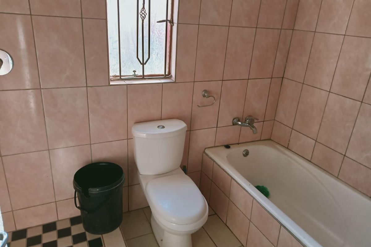 A dated bathroom featuring pink wall tiles, a standard bathtub, a toilet, and a window with security bars.