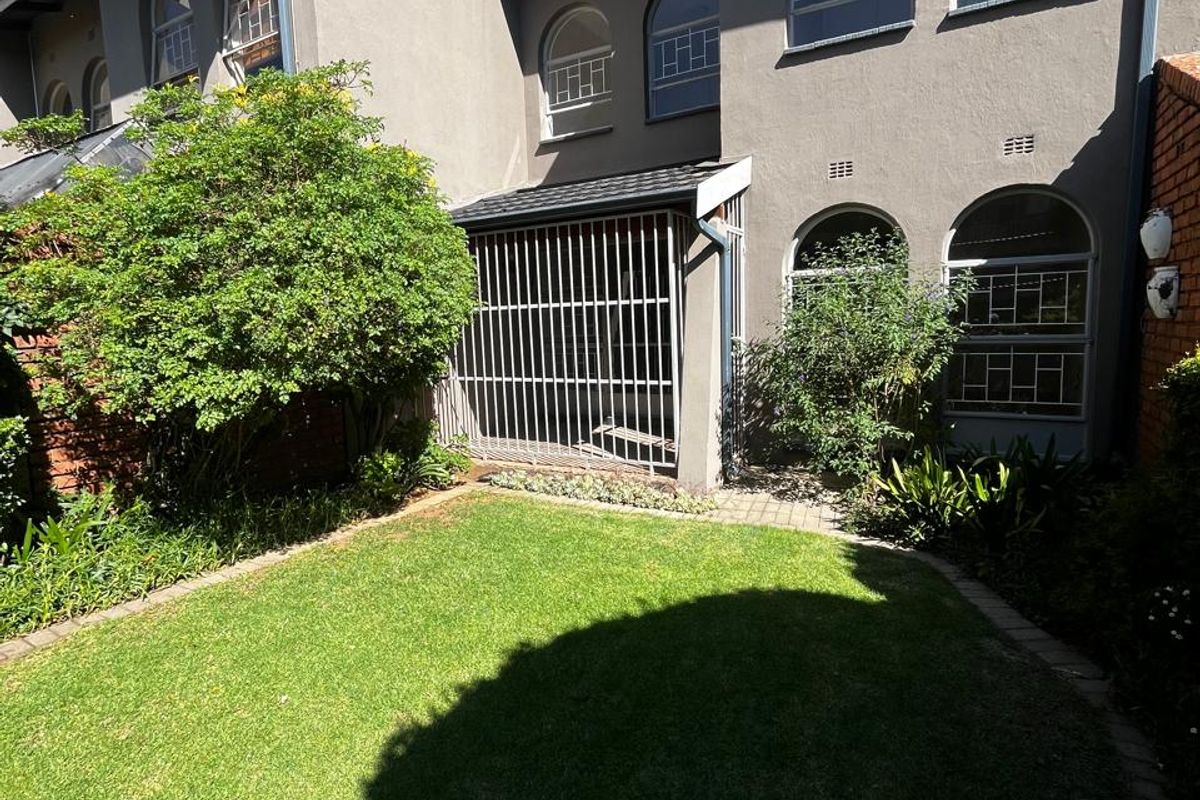 Rear view of the townhouse featuring a private garden with a lawn, paved patio area, and arched windows with security bars.