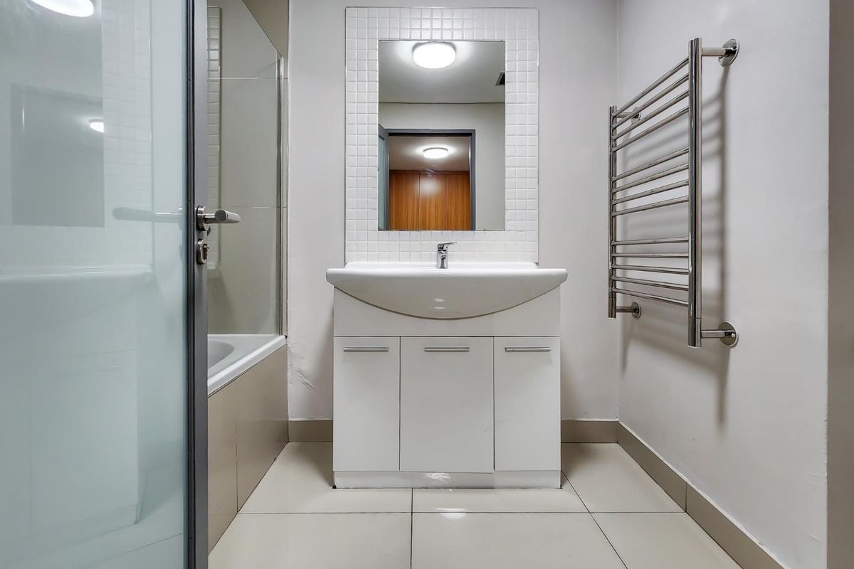 A clean, functional bathroom featuring a white vanity with basin, a glass-screened bathtub, and a wall-mounted heated towel rail.