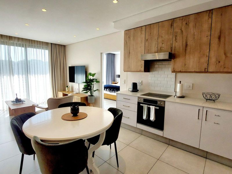 Modern open-plan living area featuring a contemporary kitchen with wood-finish cabinetry, integrated appliances, and tiled flooring, seamlessly flowing into the dining and lounge space.