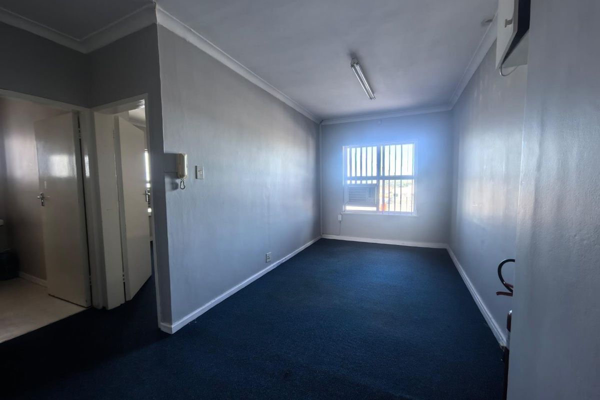An empty office space featuring grey walls, blue carpeting, security bars on the window, and a built-in air conditioning unit.