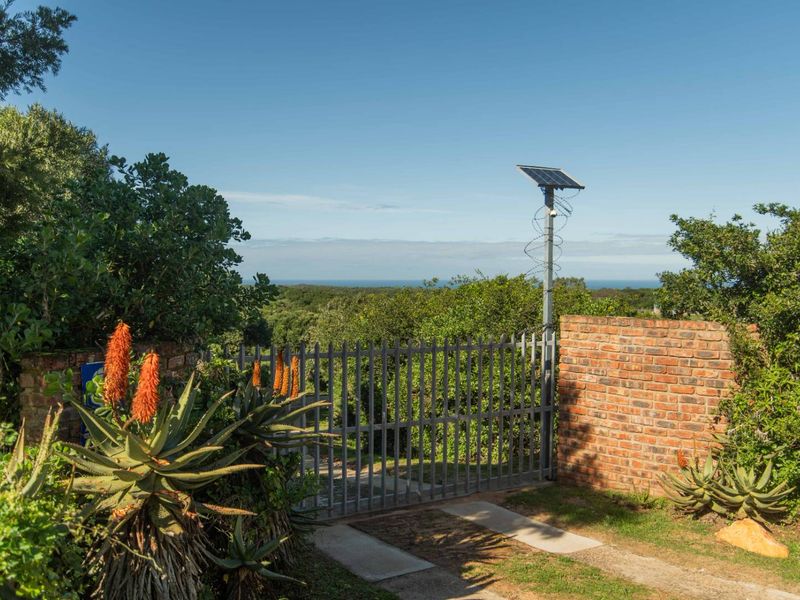 A secure front entrance featuring a metal gate, brick boundary wall, and solar-powered security system, set within a landscaped garden with coastal views.