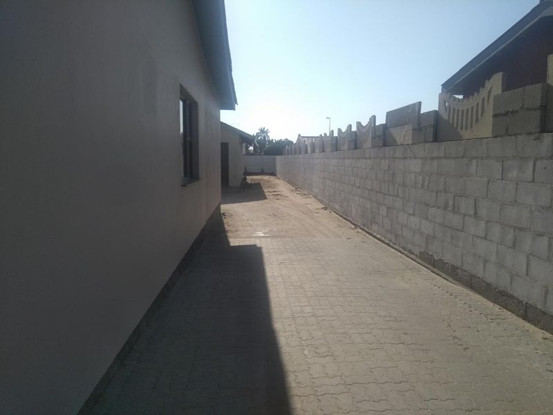 A side exterior view showing a paved driveway, a concrete block boundary wall, and an unfinished outdoor area.