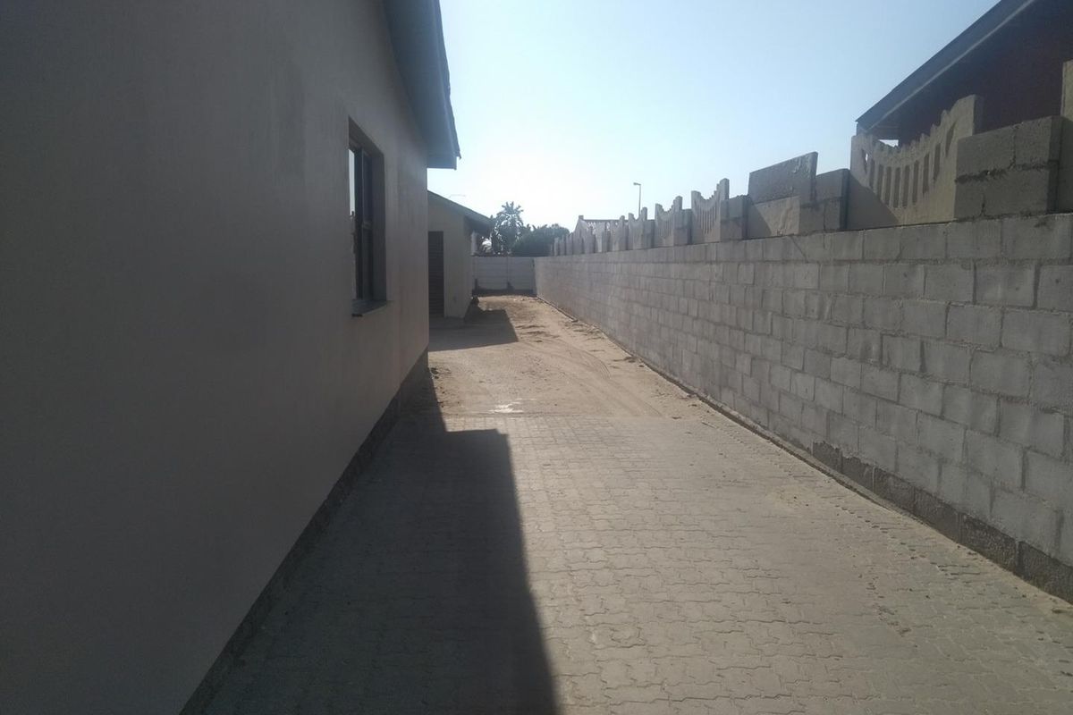 A side exterior view showing a paved driveway, a concrete block boundary wall, and an unfinished outdoor area.