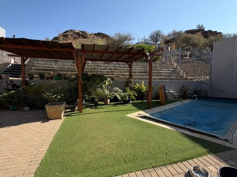 Outdoor pool area featuring a paved patio, artificial lawn, and a timber pergola, set against a terraced retaining wall and mountain backdrop.