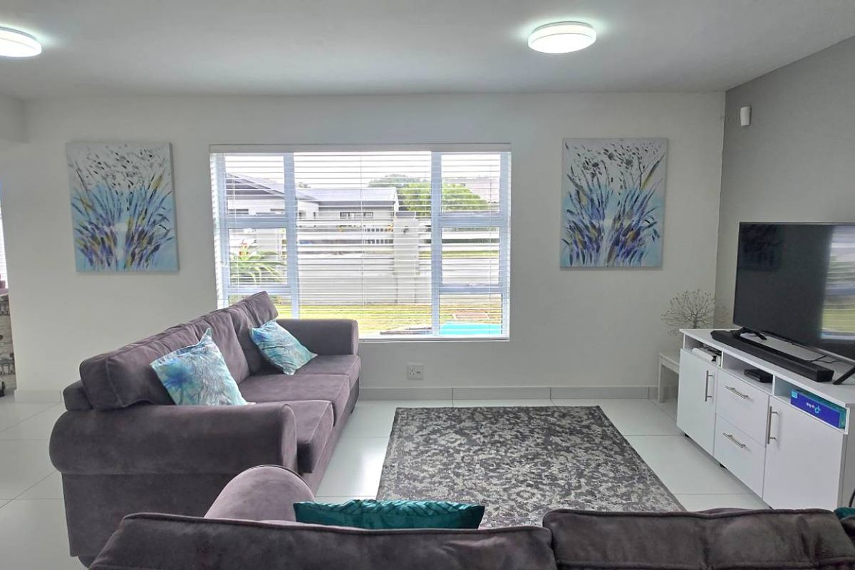 A bright and spacious living room featuring large windows with blinds, neutral tiled flooring, and recessed ceiling lighting, creating a comfortable and inviting atmosphere.