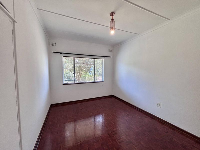 A bright, empty bedroom featuring polished parquet flooring, a built-in cupboard, and a window with fitted blinds.