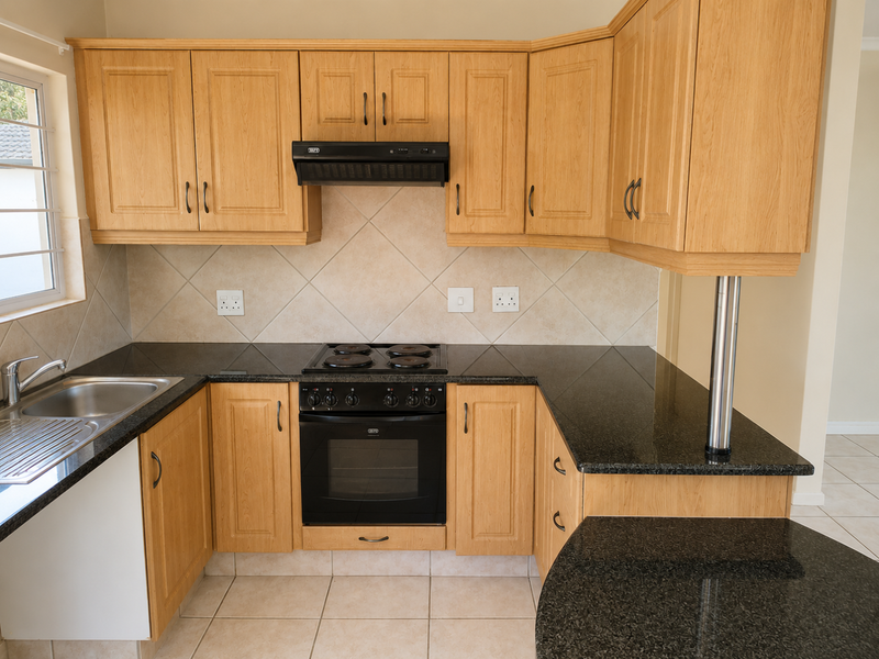 A functional kitchen featuring light wood cabinetry, dark granite countertops, a built-in oven, electric hob, and tiled finishes throughout.