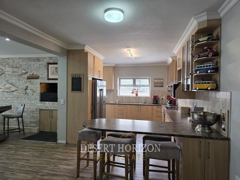 Open-plan kitchen featuring light wood cabinetry, laminate countertops, a breakfast bar, and an adjacent living area with a stone-clad built-in braai.