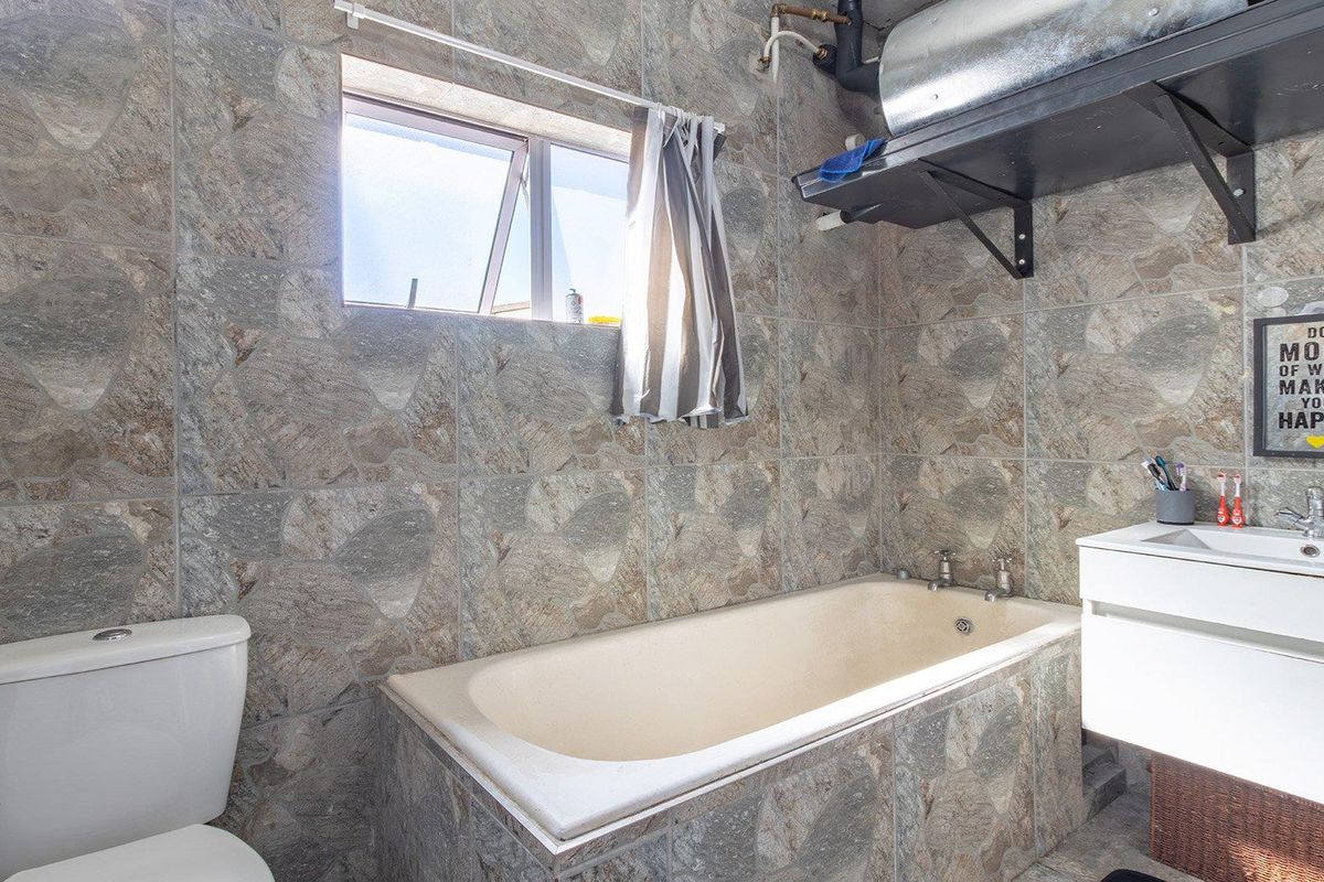 A functional bathroom featuring full-height stone-look wall tiling, a standard bathtub, a vanity basin, and a toilet, with visible exposed plumbing and geyser unit.