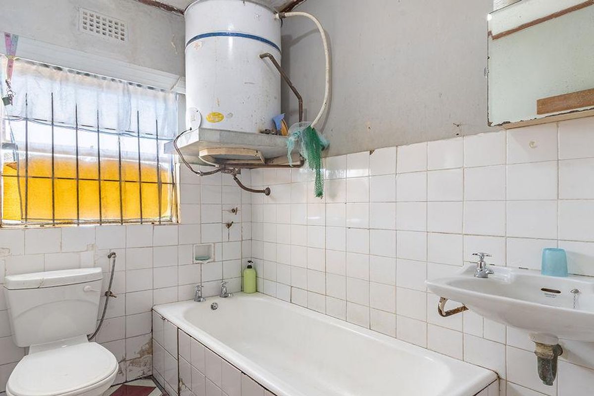 A dated bathroom featuring a bathtub, wall-mounted geyser, pedestal basin, and toilet, requiring significant renovation and modernization of finishes and fixtures.
