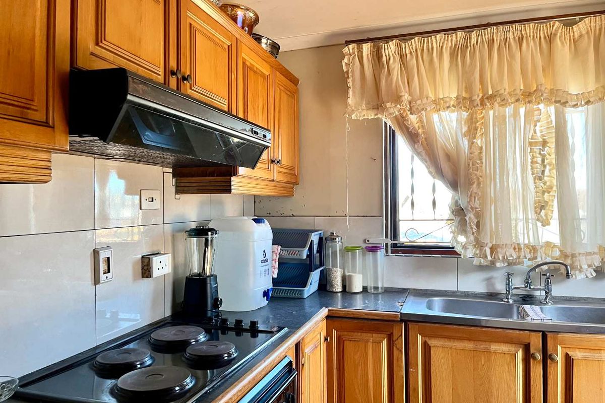 A dated kitchen featuring wooden cabinetry, an electric hob with extractor fan, and a stainless steel sink with tiled backsplash.