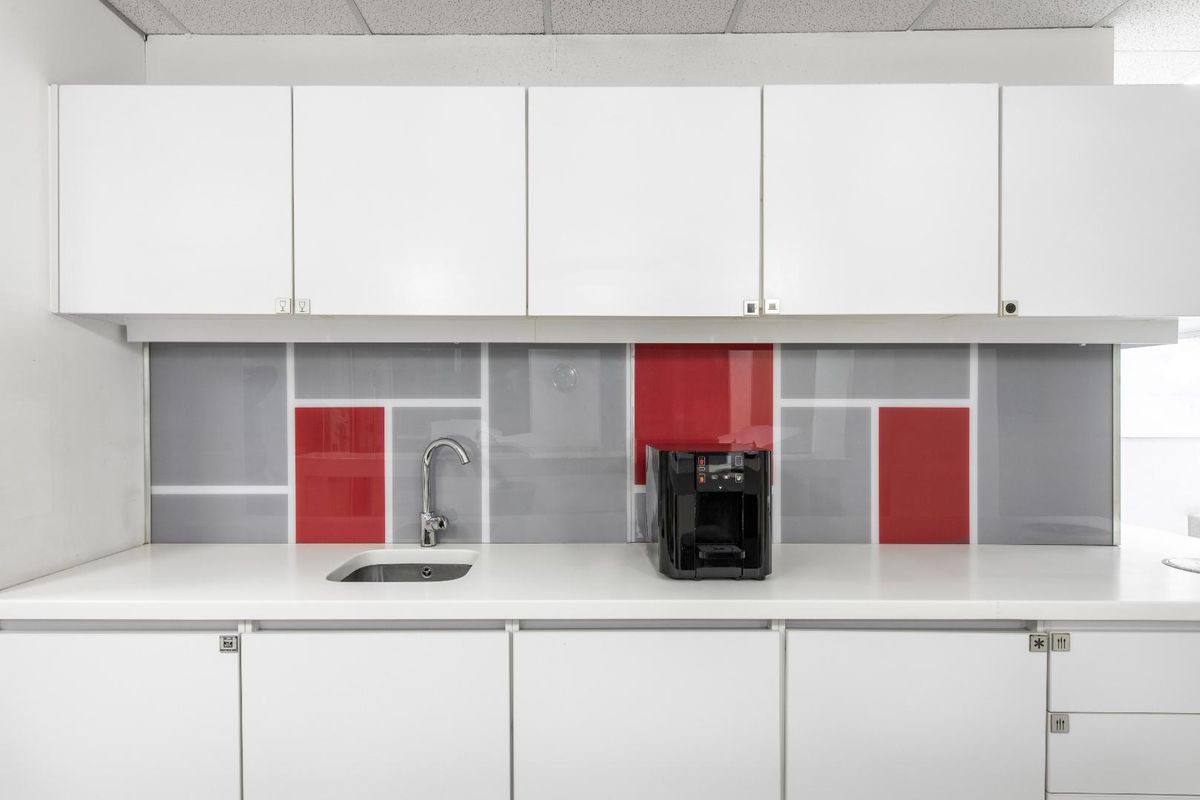 A clean office kitchenette featuring white cabinetry, a modern sink, and a contemporary red and grey glass splashback.