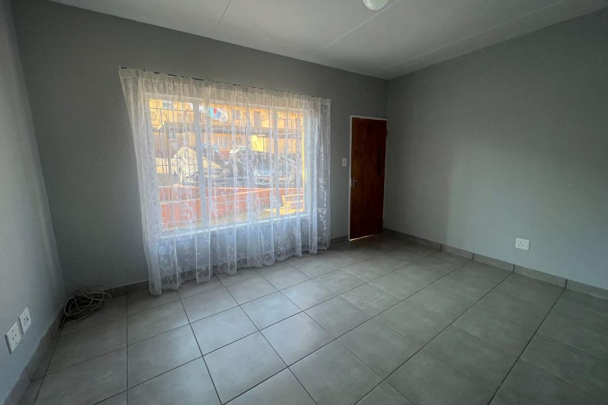 A bright, empty living room featuring neutral grey walls, large window with security bars, and durable tiled flooring throughout.