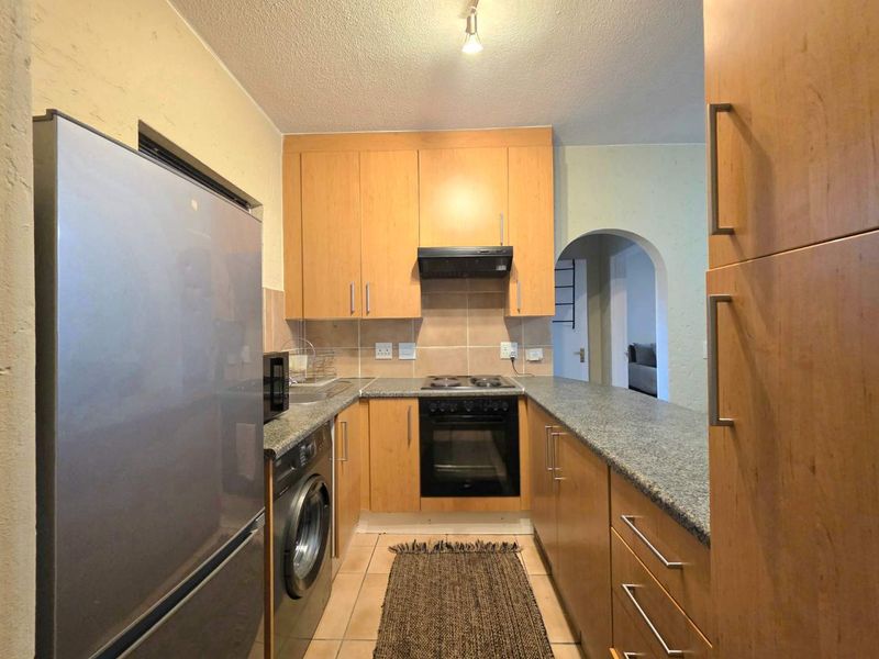 Compact kitchen featuring light wood cabinetry, granite countertops, built-in oven and hob, and tiled flooring with an arched doorway leading to the living area.
