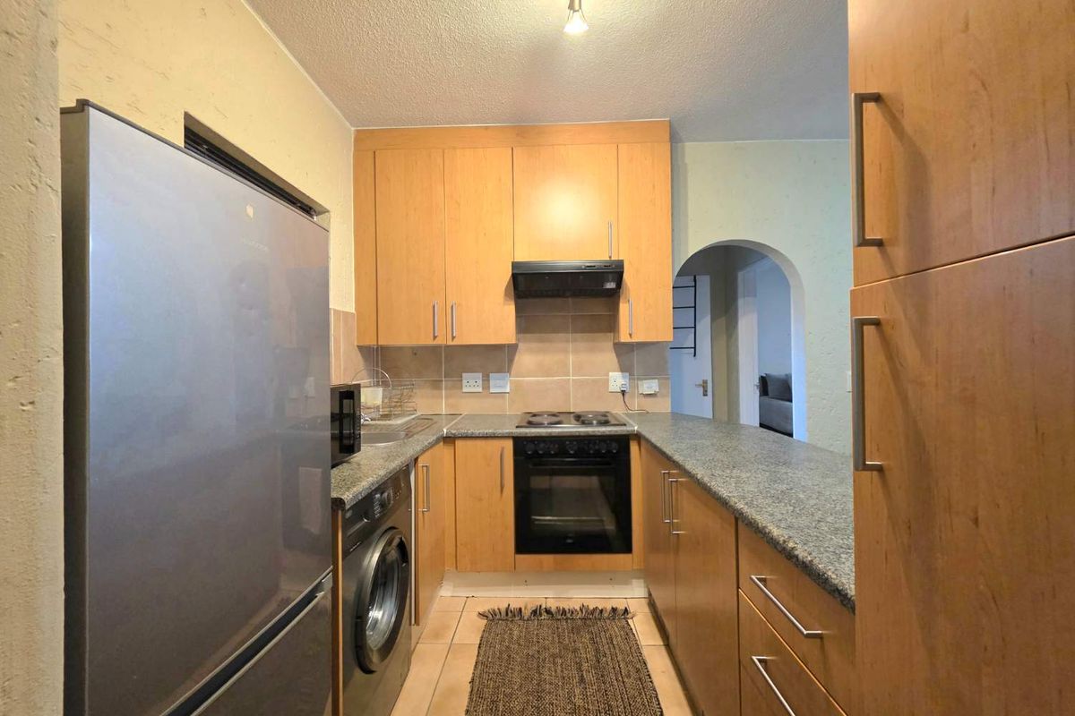 Compact kitchen featuring light wood cabinetry, granite countertops, built-in oven and hob, and tiled flooring with an arched doorway leading to the living area.