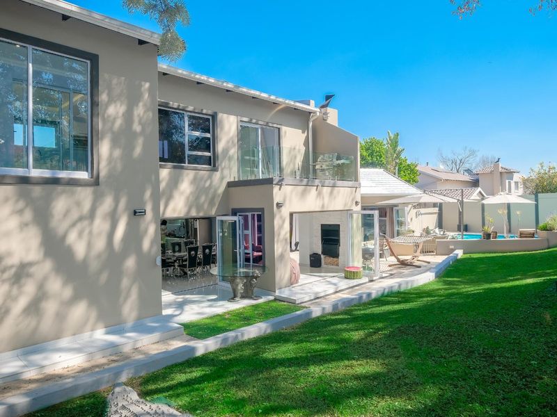 Rear view of the property featuring a landscaped garden, swimming pool, tiled patio area with built-in fireplace, and a balcony with glass balustrades.