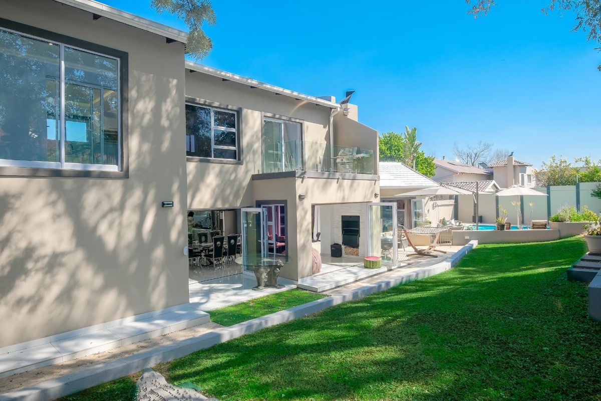Rear view of the property featuring a landscaped garden, swimming pool, tiled patio area with built-in fireplace, and a balcony with glass balustrades.
