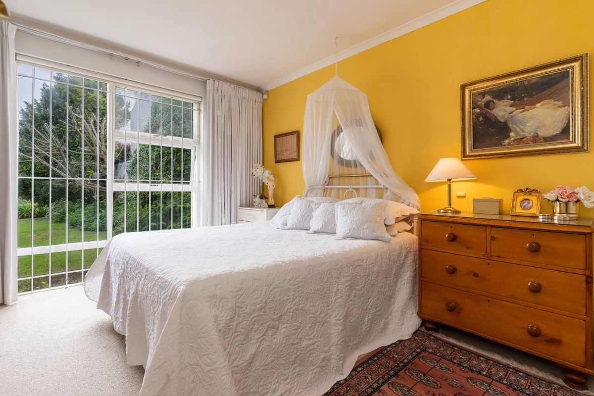 A bright bedroom featuring yellow walls, carpeted flooring, and a large window with security bars overlooking the garden.