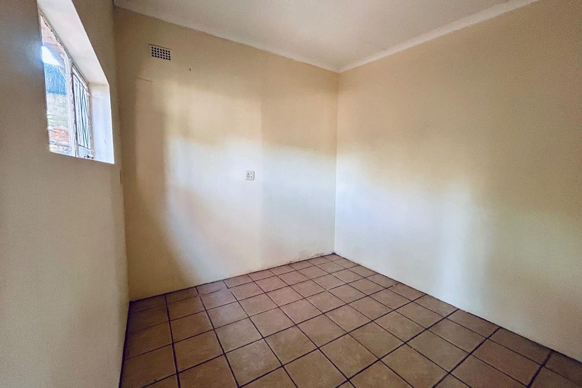 An empty, neutral-toned room featuring tiled flooring, a high-level ventilation vent, and a window with security bars.
