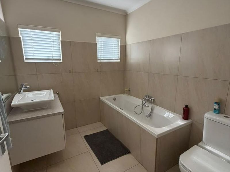 A clean and functional bathroom featuring a built-in vanity, bathtub, toilet, and neutral floor-to-ceiling wall tiling with natural light from two windows.