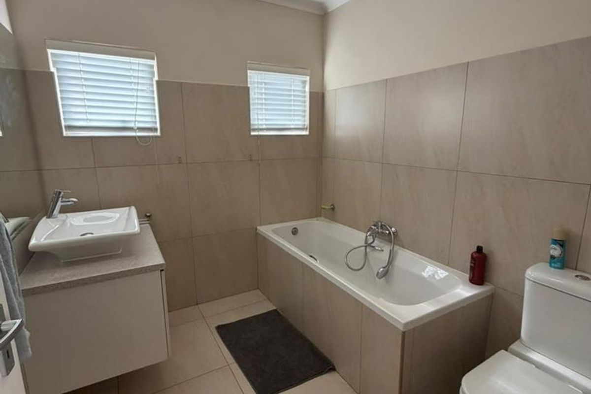 A clean and functional bathroom featuring a built-in vanity, bathtub, toilet, and neutral floor-to-ceiling wall tiling with natural light from two windows.
