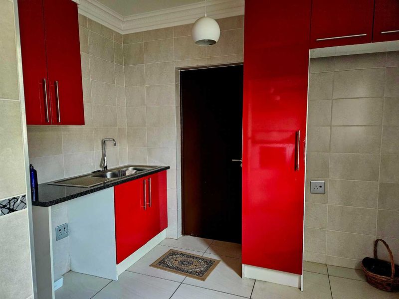 A functional scullery area featuring vibrant red cabinetry, granite countertops, a stainless steel sink, and neutral tiled finishes.