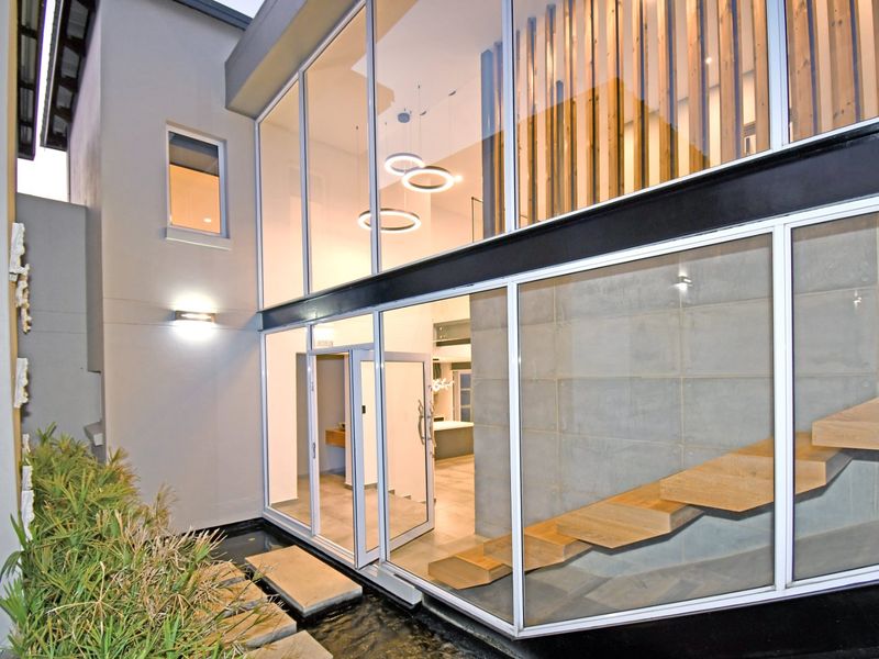 Modern architectural rear elevation featuring a double-volume glass facade, floating timber staircase, exposed concrete wall, and a tranquil water feature.