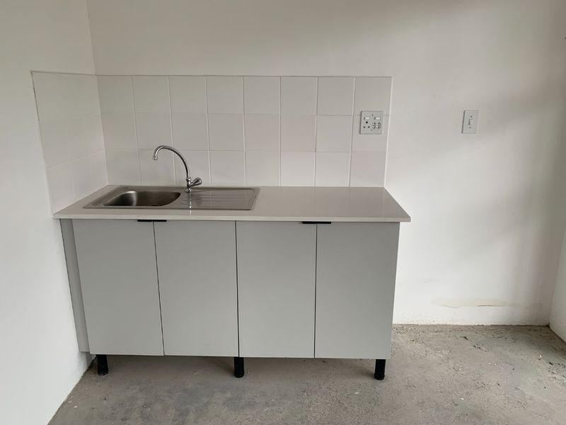 A functional laundry or utility area featuring a stainless steel sink, base cabinetry, and tiled splashback, set against a plain wall with unfinished flooring.