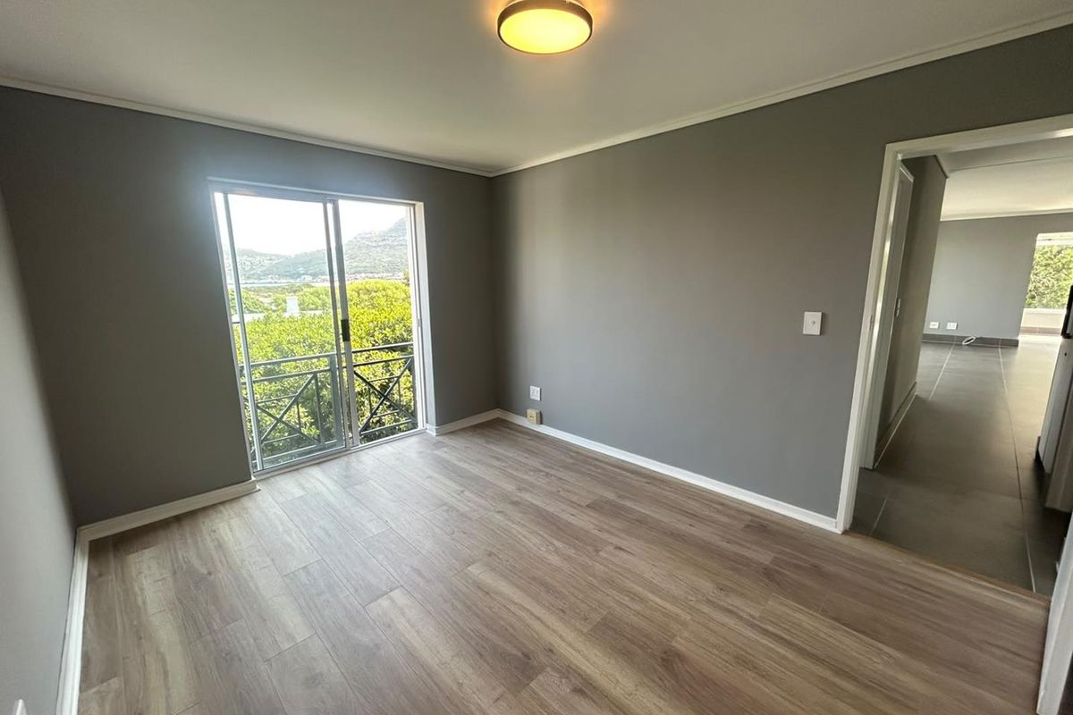 A bright, empty bedroom featuring modern laminate flooring, neutral grey walls, and a sliding glass door leading to a balcony with scenic views.