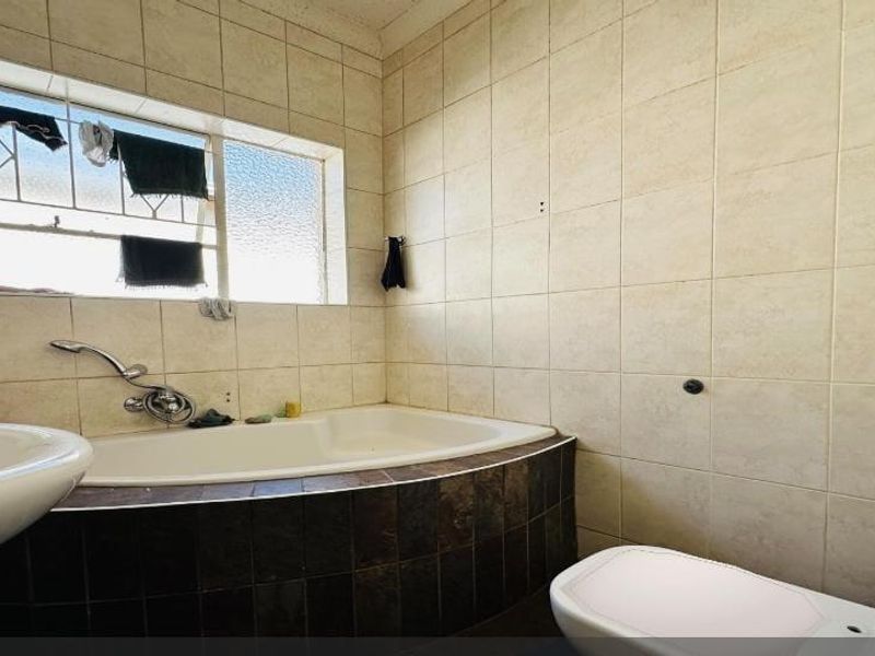 A functional bathroom featuring a built-in bathtub, pedestal basin, and toilet, finished with neutral wall tiling and security bars on the window.