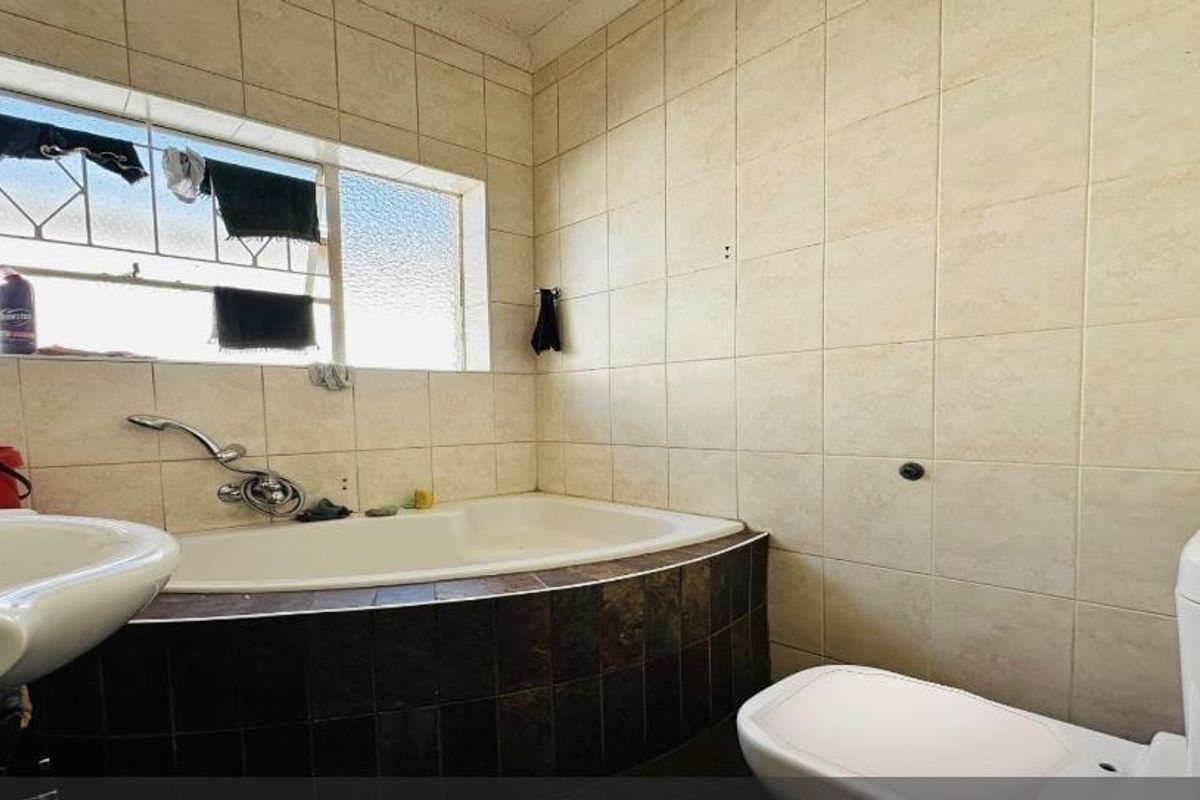 A functional bathroom featuring a built-in bathtub, pedestal basin, and toilet, finished with neutral wall tiling and security bars on the window.