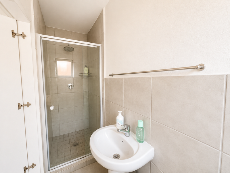 A clean and functional bathroom featuring a glass-enclosed shower, pedestal basin with mixer tap, and neutral ceramic wall tiling.