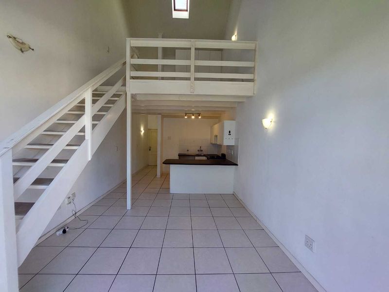 Open-plan living area featuring a mezzanine level, timber staircase, tiled flooring, and a functional kitchen with a breakfast bar.