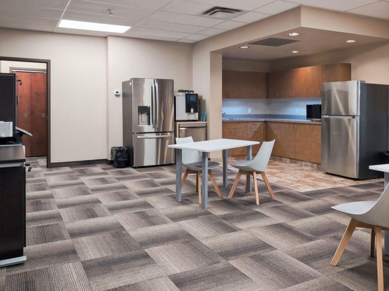An open-plan office breakroom featuring a kitchenette with built-in cabinetry, stainless steel appliances, and durable carpet tile flooring.