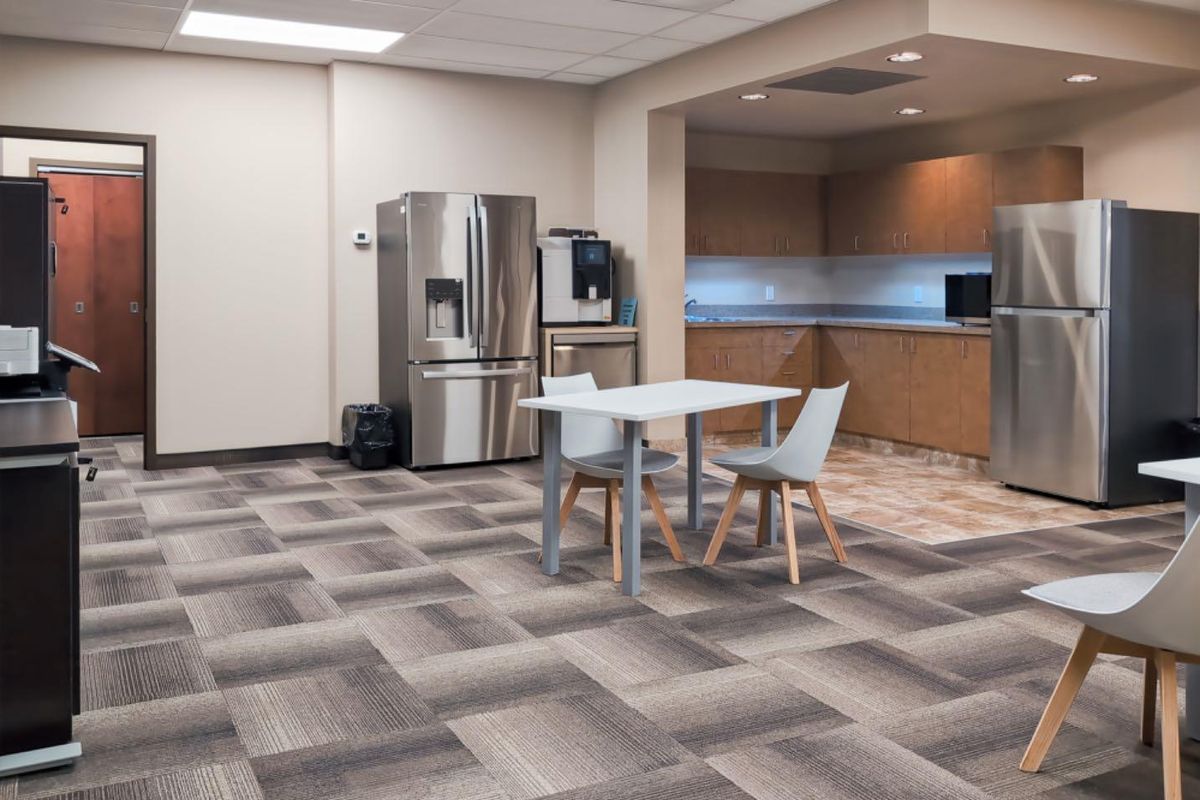 An open-plan office breakroom featuring a kitchenette with built-in cabinetry, stainless steel appliances, and durable carpet tile flooring.