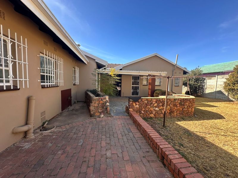 Rear exterior view featuring a brick paved patio, stone retaining walls, and a covered outdoor area with security bars on windows.