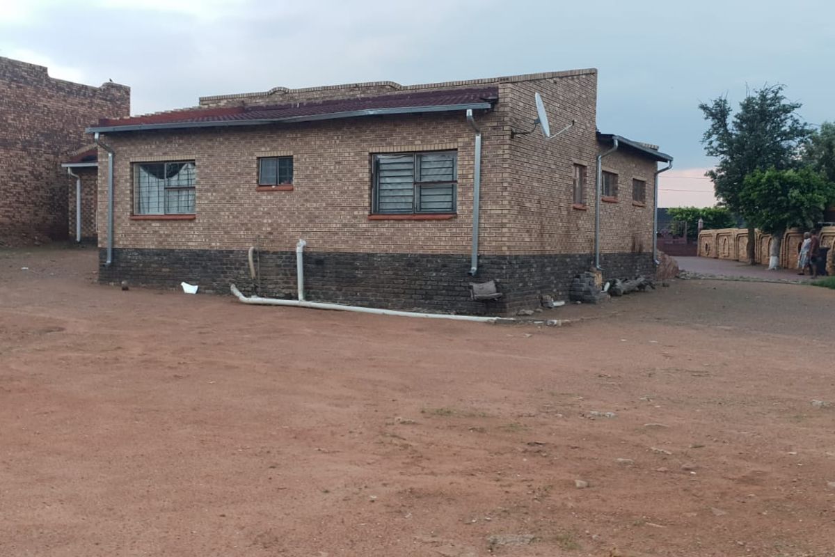 Exterior view of a single-story brick house featuring metal window frames and a corrugated metal roof, situated on an unpaved lot.