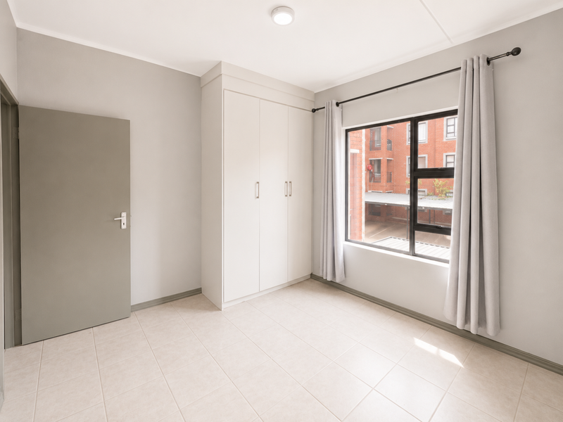 A bright, empty bedroom featuring neutral tiled flooring, built-in cupboards, and a large window providing natural light.