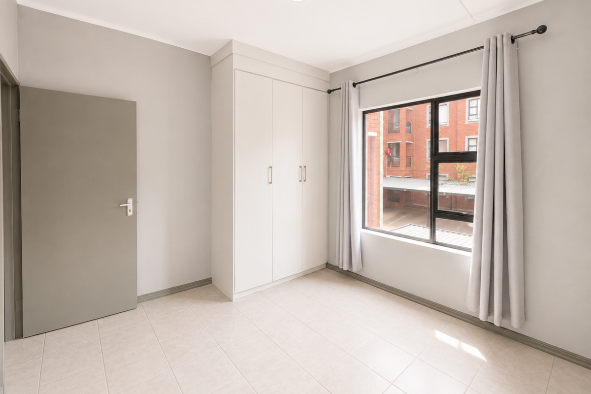 A bright, empty bedroom featuring neutral tiled flooring, built-in cupboards, and a large window providing natural light.