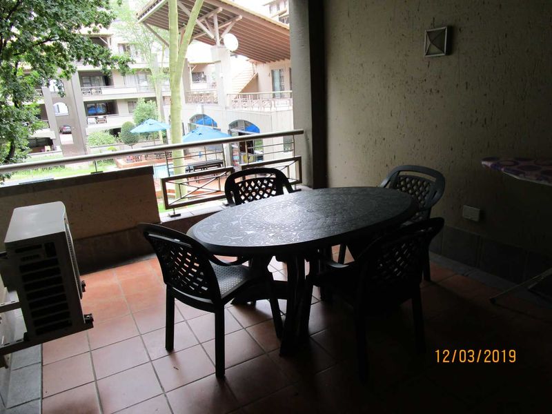 A covered balcony featuring tiled flooring, a metal balustrade, and an external wall light, overlooking the apartment complex grounds.