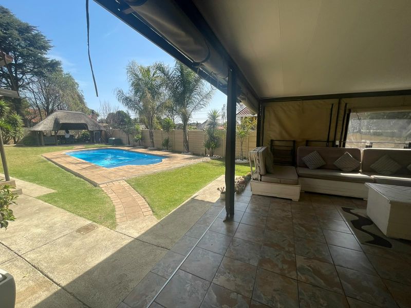 Spacious covered patio overlooking a well-maintained garden, swimming pool, and a traditional thatched lapa, perfect for outdoor entertaining.
