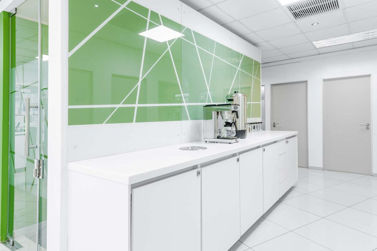 Modern office kitchenette featuring sleek white cabinetry, integrated sink, and a vibrant green glass splashback with geometric detailing.