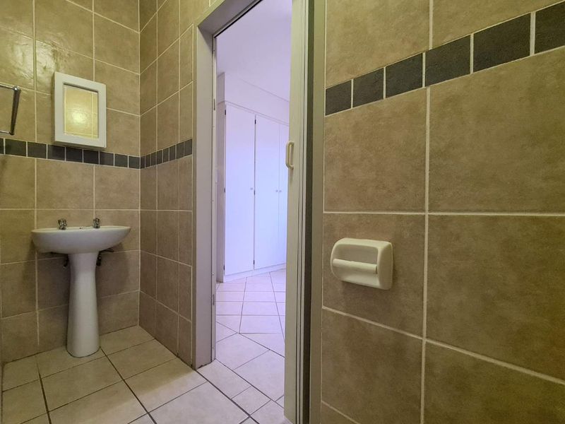 A functional bathroom featuring neutral ceramic wall and floor tiles, a pedestal basin, and a doorway leading to an adjacent room with built-in cupboards.