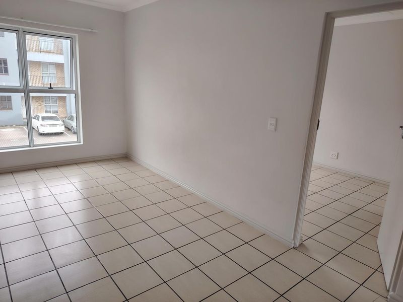 A clean, empty bedroom featuring neutral tiled flooring, white walls, and a large window providing natural light and views of the apartment complex parking area.