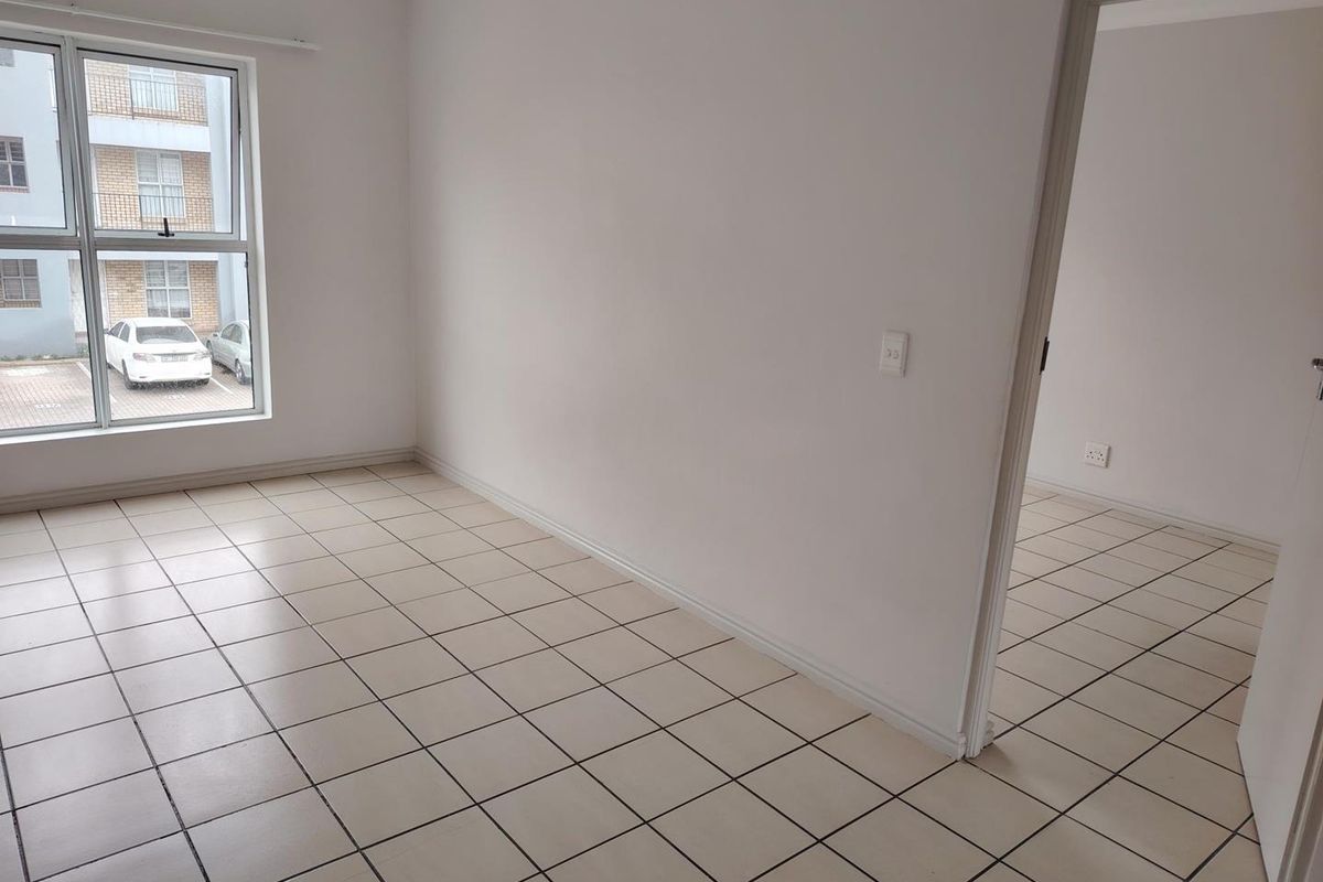 A clean, empty bedroom featuring neutral tiled flooring, white walls, and a large window providing natural light and views of the apartment complex parking area.