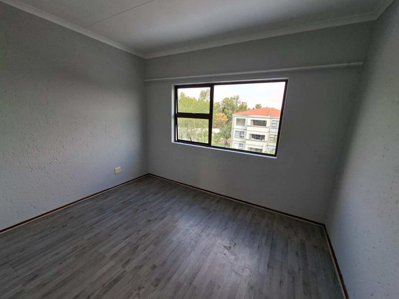 An empty bedroom featuring grey laminate flooring, textured walls, and an aluminium-framed window with a view of the surrounding apartment complex.
