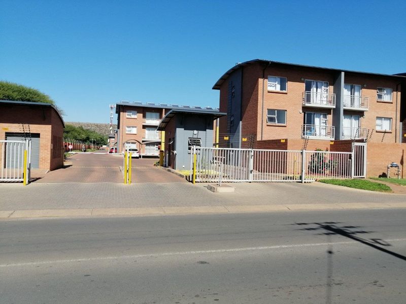 Front view of the apartment complex entrance featuring a secure gated access point, guard house, and multi-story brick residential buildings.
