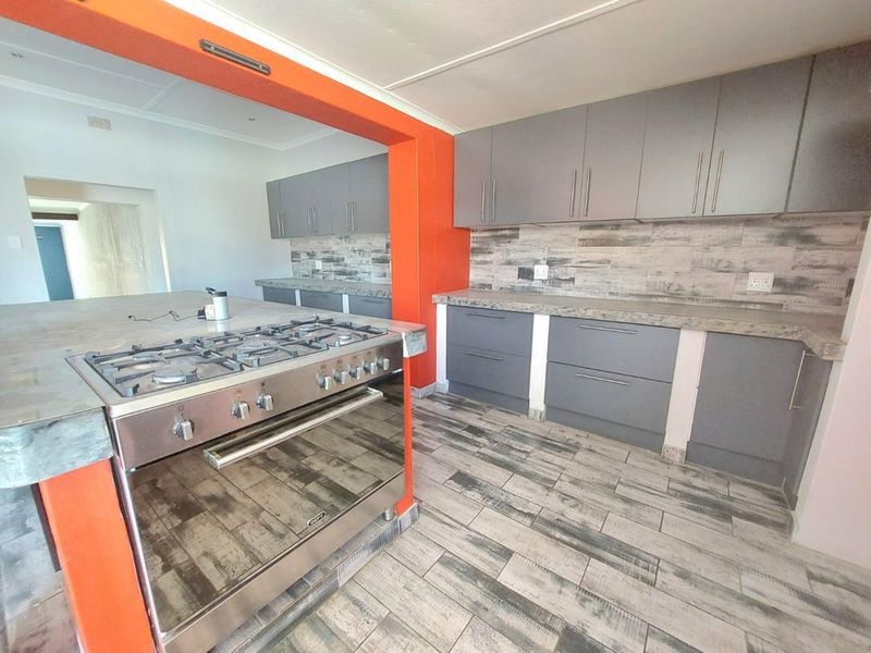 Modern kitchen featuring grey built-in cabinetry, stone countertops, a gas stove with oven, and wood-look tiled flooring with a bold orange accent wall.