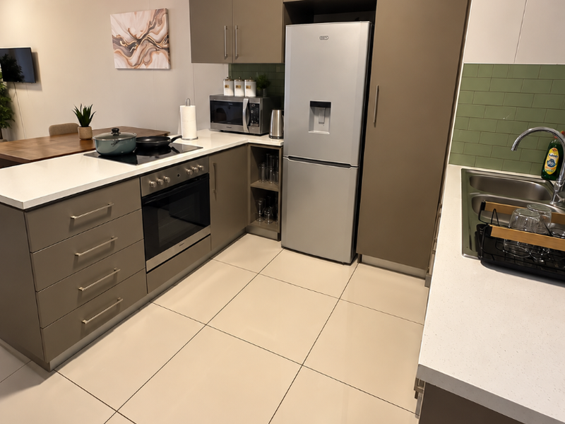 Modern kitchen featuring sleek taupe cabinetry, white stone countertops, a tiled backsplash, and integrated appliances, seamlessly flowing into the adjacent living area.